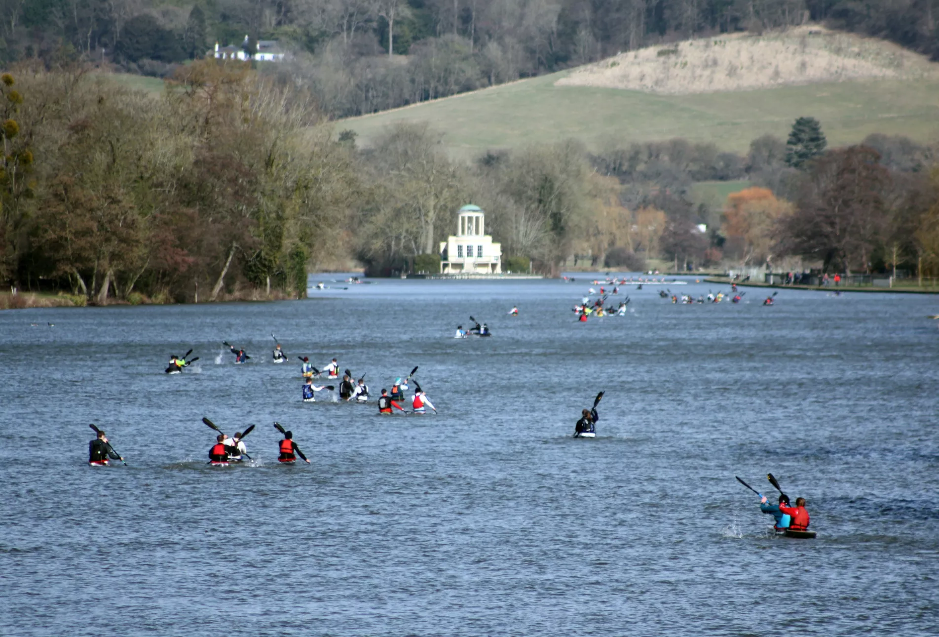 Wide view of a river filled with dozens of kayakers paddling along the water. Tree-lined banks and rolling hills surround the river, with a pale-coloured pavilion visible in the distance under a bright, overcast sky.