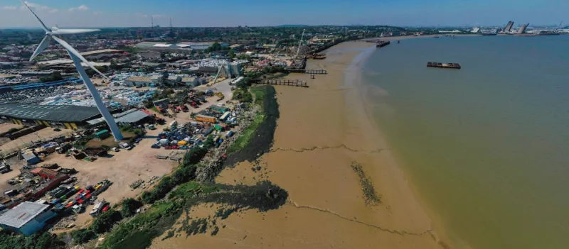 Aerial view of an industrial waterfront area with a large wind turbine, warehouses, and scattered machinery on land beside a wide, muddy shoreline. The river stretches across the right side of the image with several barges and piers visible. Urban buildings and infrastructure extend into the background under a clear sky